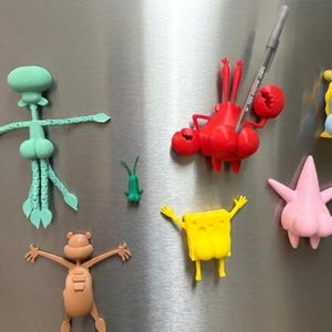 May include: A collection of colorful cartoon character magnets, including Squidward, Mr. Krabs, SpongeBob, Patrick Star, and others. A red Mr. Krabs magnet holds a pen. The magnets are attached to a silver refrigerator.