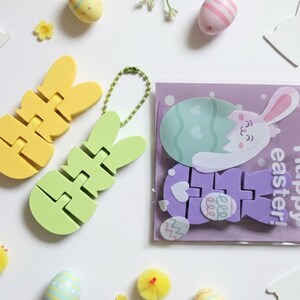 May include: An Easter-themed flat lay featuring yellow and green bunny-shaped puzzles, decorative eggs, and a purple card with a bunny and the text "Happy Easter!". The scene is set on a white surface with floral and chick accents.