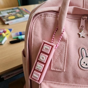 May include: A pink backpack with a matching keychain spelling "LORI" in white letters. The backpack has a bunny patch and a star zipper pull. The scene suggests a school or student setting.