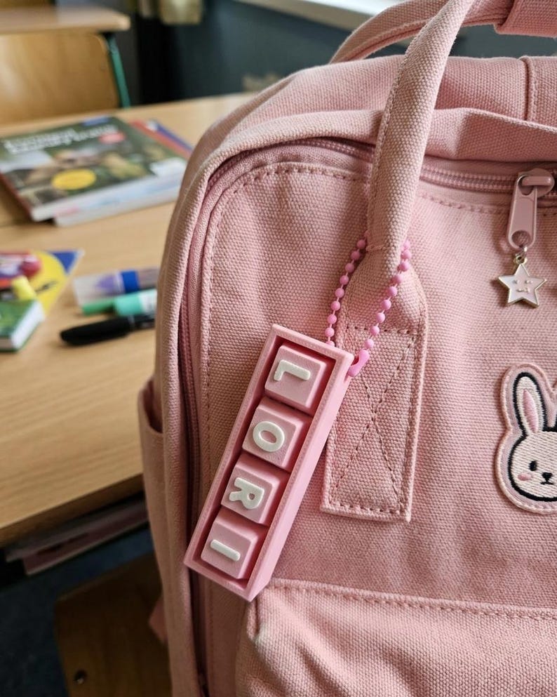 May include: A pink backpack with a matching keychain spelling "LORI" in white letters. The backpack has a bunny patch and a star zipper pull. The scene suggests a school or student setting.