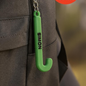 May include: A green hockey stick-shaped keychain with the name "SIMON" in black lettering. It's attached to a silver chain, set against a dark grey fabric. A red circle with "Made to Order" is visible. The keychain is a personalised accessory.