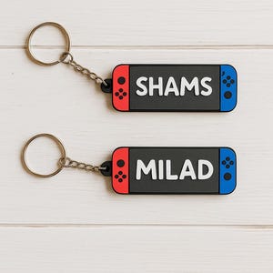 May include: Two keyrings shaped like game controllers. One keyring has the name "SHAMS" in white letters on a black background, with red and blue accents. The other keyring has the name "MILAD". Both have silver key rings.