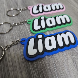 Puede incluir: Llaveros con el nombre "Liam" en blanco, contorneado en negro, sobre fondos rosa, verde y azul. Cada llavero tiene una cadena y un anillo plateados. Los llaveros están sobre una superficie de madera.
