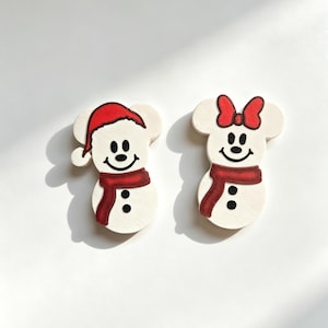 May include: Two white snowman-shaped ornaments with mouse-like ears. The left figure wears a red Santa hat and a red scarf. The right figure wears a red bow and a red scarf. Both have black eyes, a black smile, and two black buttons on their bodies.