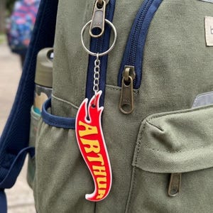 May include: A flame-shaped keychain attached to a green backpack zipper. The keychain features a red border with a white outline and a yellow center containing the name ARTHUR in a bold, stylized font, suspended by a silver metal ring and chain.