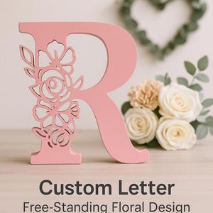May include: A pink wooden letter 'R' with a floral design cutout on the left side. The letter is free-standing and has a smooth finish. The text at the bottom reads 'Custom Letter Free-Standing Floral Design'.