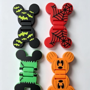 May include: Four hinged plastic clips shaped like mouse heads with Halloween designs. One is black with yellow bats, one is red with black spiderwebs, one is green with black stitches, and one is orange with black pumpkin face details.