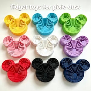 May include: Nine silicone fidget toys shaped like mouse heads with concentric circular patterns, arranged in a grid. The colors include yellow, light blue, green, pink, white, purple, red, black, and dark blue. Text reads: fidget toys for pixie dust.