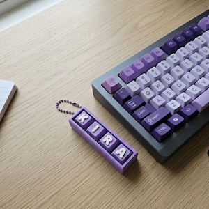May include: A custom keyboard with purple and white keycaps sits on a wooden desk. A matching keychain spells out "KIRA" in white letters on a purple background. The keyboard has a dark gray frame.