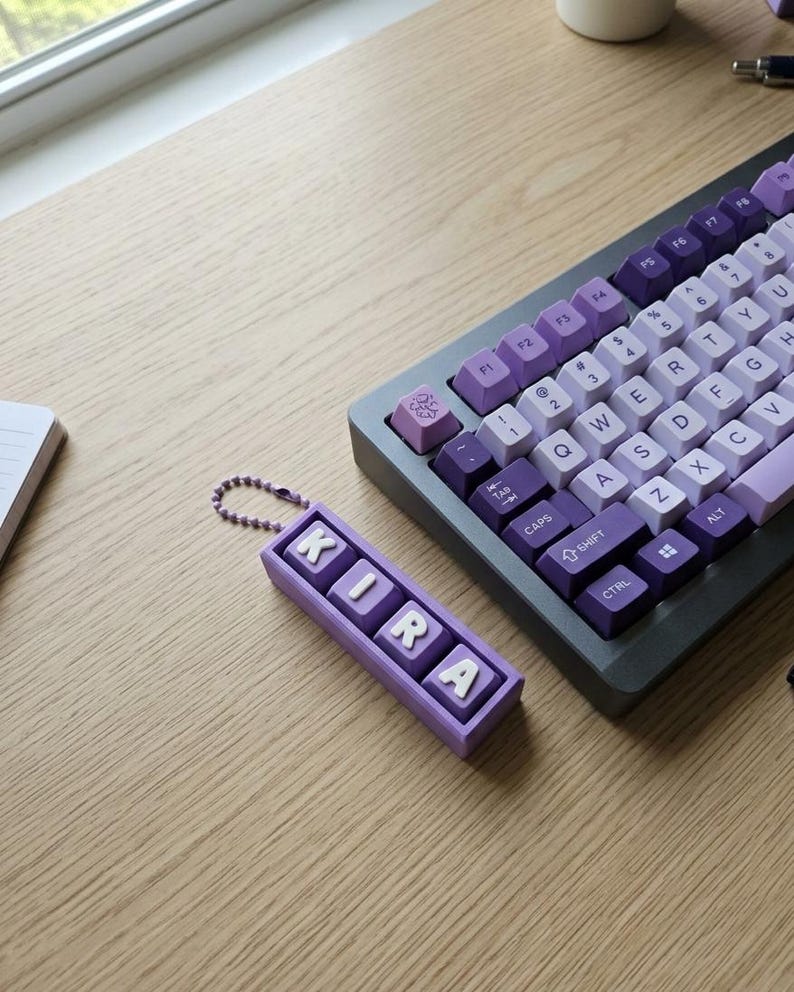 May include: A custom keyboard with purple and white keycaps sits on a wooden desk. A matching keychain spells out "KIRA" in white letters on a purple background. The keyboard has a dark gray frame.