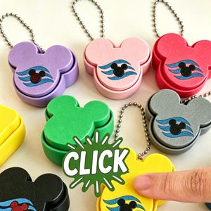 May include: A selection of Mickey Mouse-shaped keyrings in various colours. Each keyring has a two-tone design with a black Mickey Mouse head and blue wave accents. The word "CLICK" is displayed on a green burst graphic.
