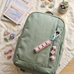 May include: A sage green backpack with a pink keychain spelling "MONA". The backpack has a front zipper pocket and a small patch with a mountain design. Several small pink cubes and a succulent plant in a gray pot are also visible.