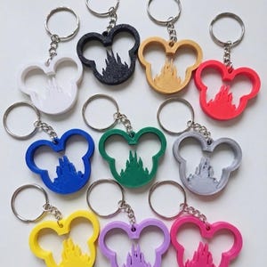 May include: A selection of Mickey Mouse head-shaped keyrings in various colours, each with a castle silhouette. Keyrings are in white, black, yellow, red, blue, green, grey, purple, and pink. Each keyring has a silver keyring attached.