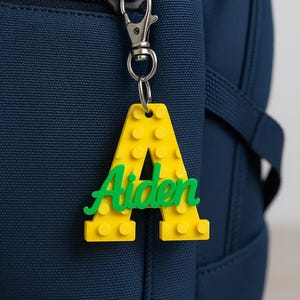 以下が含まれることがあります： 黄色の「A」の形をしたキーホルダー。緑色の筆記体で「Aiden」と書かれています。キーホルダーはシルバーの留め金とリングに付けられており、濃いネイビーブルーのバックパックにぶら下がっています。