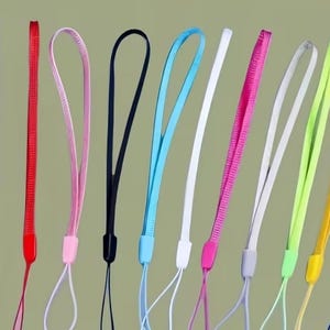 May include: A collection of colorful wrist lanyards in various shades, including orange, purple, red, pink, black, blue, white, and yellow. Each lanyard features a loop for the wrist and a thin cord for attaching to a device.
