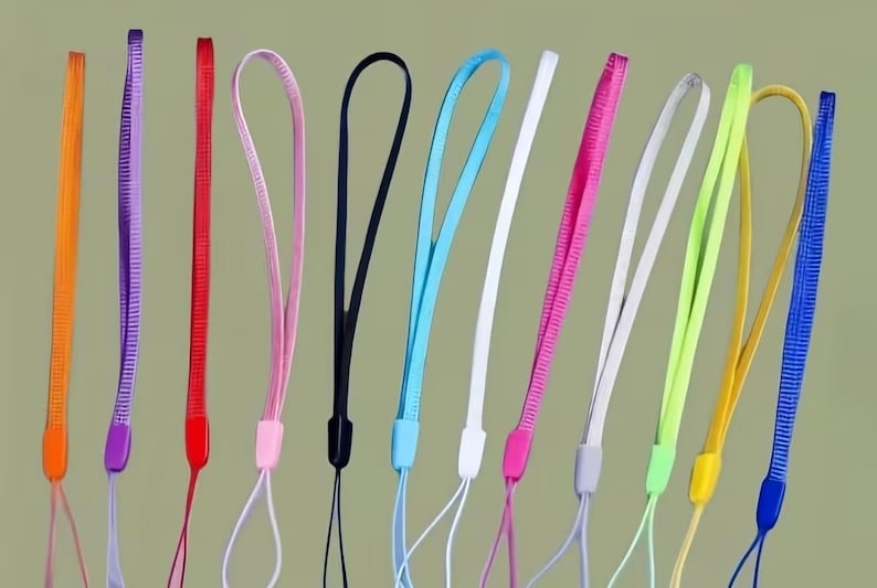 May include: A collection of colorful wrist lanyards in various shades, including orange, purple, red, pink, black, blue, white, and yellow. Each lanyard features a loop for the wrist and a thin cord for attaching to a device.