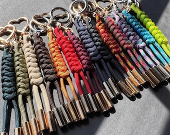 May include: A collection of paracord keychains featuring braided designs in various solid colors and multi-colored patterns. Each keychain is attached to a silver-toned heart-shaped ring and finished with two metallic cylindrical end caps.