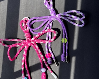 Puede incluir: Dos llaveros hechos a mano con paracord. Uno es rosa y blanco, el otro morado y blanco. Cada llavero presenta un diseño en forma de lazo con cuentas decorativas y puntas metálicas. Los llaveros se muestran sobre una superficie gris oscuro.