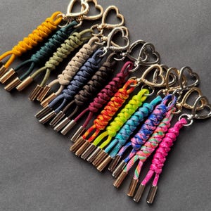 May include: A collection of colorful paracord keychains with heart-shaped clasps. The keychains feature a variety of colors, including yellow, green, blue, and pink, with metallic gold and silver accents. The braided design adds texture and visual interest.