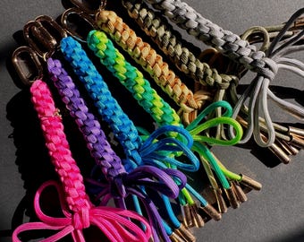 Puede incluir: Una colección de llaveros de paracord coloridos. Cada llavero presenta un diseño trenzado en diferentes colores, incluyendo rosa, morado, azul, verde, amarillo y gris. Los llaveros tienen cierres dorados y borlas decorativas.