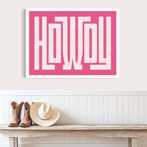 Op de afbeelding: Een rechthoekige wandkunstprint met het woord HOWDY in grote, dikke, witte blokletters tegen een levendige roze achtergrond. Het kunstwerk hangt boven een houten bankje met twee bruine leren cowboylaarzen en een beige cowboyhoed.