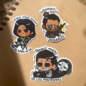 May include: Four colourful stickers featuring cartoon characters with text. The first sticker shows a cartoon woman with brown hair and a green shirt, sitting on a chair. The text reads "Even dogs know not to bark at me". The second sticker shows a cartoon man with brown hair and a black shirt, holding a gun. The text reads "Weapons hot vaqueros!". The third sticker shows a cartoon man with brown hair and a black shirt, crying. The text reads "Tengo miedo de los fantasmas". The fourth sticker shows a cartoon skull with a black and white shirt.