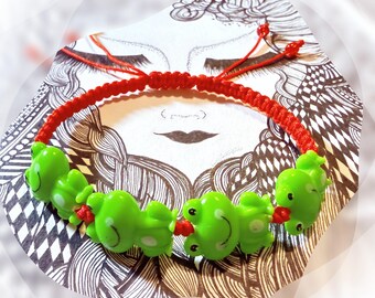 May include: A handmade bracelet featuring bright green frog charms strung on a red braided cord. The bracelet is set against a black and white illustration of a face.
