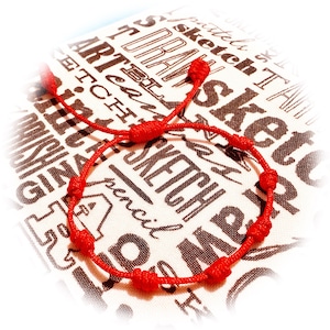 May include: A red string bracelet with multiple knots. The bracelet is adjustable and rests on a white and brown patterned background with the words "ART", "SKETCH", and "DRAW" printed on it.