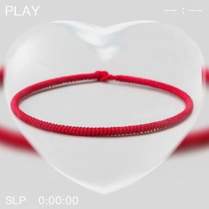 Può includere: Un braccialetto rosso brillante, annodato a un'estremità, è mostrato su uno sfondo morbido e sfocato. Il braccialetto è un anello continuo di materiale intrecciato. La parola "PLAY" è visibile nell'angolo in alto a sinistra.