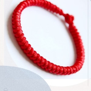 Peut inclure: Un bracelet tressé rouge vif, présentant un design en boucle continue. Le bracelet est fait d'un matériau tissé serré, créant une surface texturée. La couleur rouge vif est uniforme sur tout le bracelet, sur fond blanc.