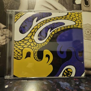 Peut inclure: Un boîtier de CD avec un design abstrait coloré. L'œuvre présente des formes jaunes, bleues et blanches sur fond noir. Le design a une ambiance psychédélique avec des formes organiques et fluides. Le boîtier est en plastique transparent.