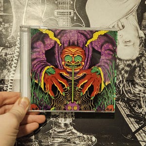 Könnte beinhalten: Ein CD-Gehäuse mit einer farbenfrohen Illustration eines grünhäutigen, rotkralltigen Wesens mit einer langen Nase und einem violetten Gewand. Das Wesen ist von grünen Pflanzen und einem wirbelnden Muster umgeben.