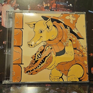 Pode incluir: Caixa de CD com uma ilustração de desenho animado de um porco montando um jacaré, ambos em armadura. O porco usa um chapéu floral. A obra de arte é em tons de amarelo, laranja e marrom. A caixa do CD é transparente.