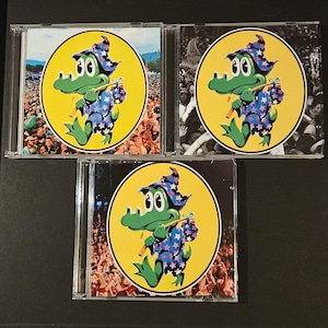 May include: Three CD cases featuring a cartoon alligator character in a wizard hat and robe, set against a yellow circle. The alligator is green with a blue and white star-patterned robe. The background of each case shows a crowd of people.