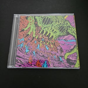 Puede incluir: Una caja de CD con una ilustración vibrante y surrealista. La obra de arte representa una gran forma orgánica verde que domina la parte superior, con figuras de varios colores debajo. El fondo es una mezcla de morado, naranja y rosa.