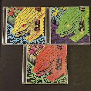May include: Three CD cases with colorful artwork. Each case features a stylized alligator head with a railway track, water, and floral elements. The color schemes are yellow, green, and red, with black and blue backgrounds. The number "00" is visible on each case.