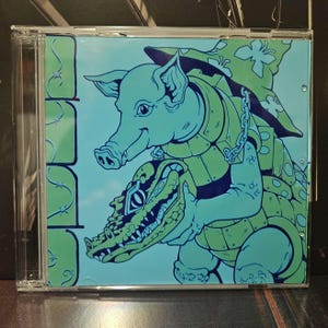 Peut inclure: Boîtier de CD avec une illustration de dessin animé d'un cochon et d'un alligator dans des tons de vert et de bleu. Le cochon est au-dessus de l'alligator, tous deux portant une armure. Le fond est bleu clair.