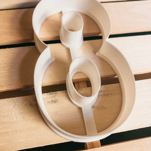 May include: White plastic cookie cutter shaped like the number eight. The cutter is on a wooden surface.