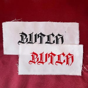 May include: Two white fabric patches with the word "BUTCH" printed in a gothic font. One patch has black text, and the other has red text. The patches are on a red background.