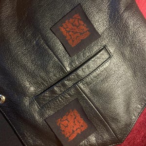 May include: Black leather vest with two square patches featuring a reddish-brown floral design. The vest has a pocket and metal snap closures. The leather has a textured appearance.