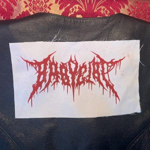 Könnte beinhalten: Ein weißes Stoffabzeichen mit dem roten Text "BABYGIRL" in einer gotischen Schrift ist auf eine schwarze Lederjacke genäht. Das Abzeichen hat ein verwittertes, tropfendes Design.