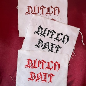 May include: Three white fabric patches with the words "BUTCH" and "BUTCH BAIT" printed in a gothic font. The text is in red and black ink. The patches are arranged on a red background.