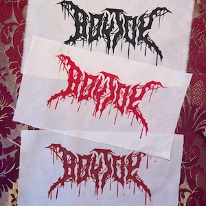 May include: Three fabric patches with the word "BODHI" in a gothic font. The top patch has black text, the middle patch has red text, and the bottom patch has red text. The patches are on a patterned red and gold background.