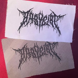 May include: Two patches with the word "BABYGRRL" in a black, gothic-style font. One patch is on white fabric, the other on beige fabric. The patches are displayed on a red surface.