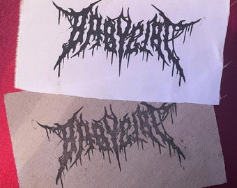 May include: Two patches with the word "BABYGRRL" in a black, gothic-style font. One patch is on white fabric, the other on beige fabric. The patches are displayed on a red surface.