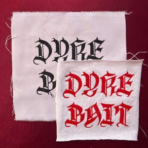 May include: Two white fabric patches with black and red gothic lettering. The top patch reads "DYKE" in black. The bottom patch reads "DYKE BAIT" in red. The patches are on a red background.