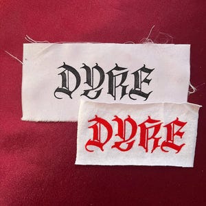 Op de afbeelding: Twee witte stoffen labels met het woord "DYRE" gedrukt in een gotisch lettertype. Eén label heeft zwarte tekst en de andere rode tekst. De labels staan op een bordeauxrode achtergrond.