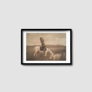 Könnte beinhalten: Ein Schwarzweißfoto eines Indianers in traditioneller Kleidung, der auf einem weißen Pferd reitet. Das Pferd trinkt aus einem kleinen Teich in einem grasbewachsenen Feld.