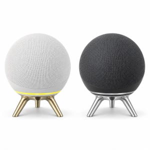 May include: Two spherical smart speakers with fabric mesh covers, one white and one charcoal gray. Each speaker is mounted on a three-legged metal stand. The white speaker features a gold-toned base with a glowing yellow light ring.