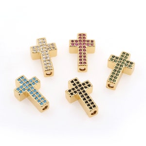 May include: Five gold cross charms with different colored crystals. The crosses are all the same size and shape, and they are all set with small, round crystals. The crystals are in the colors of white, red, green, blue, and black.